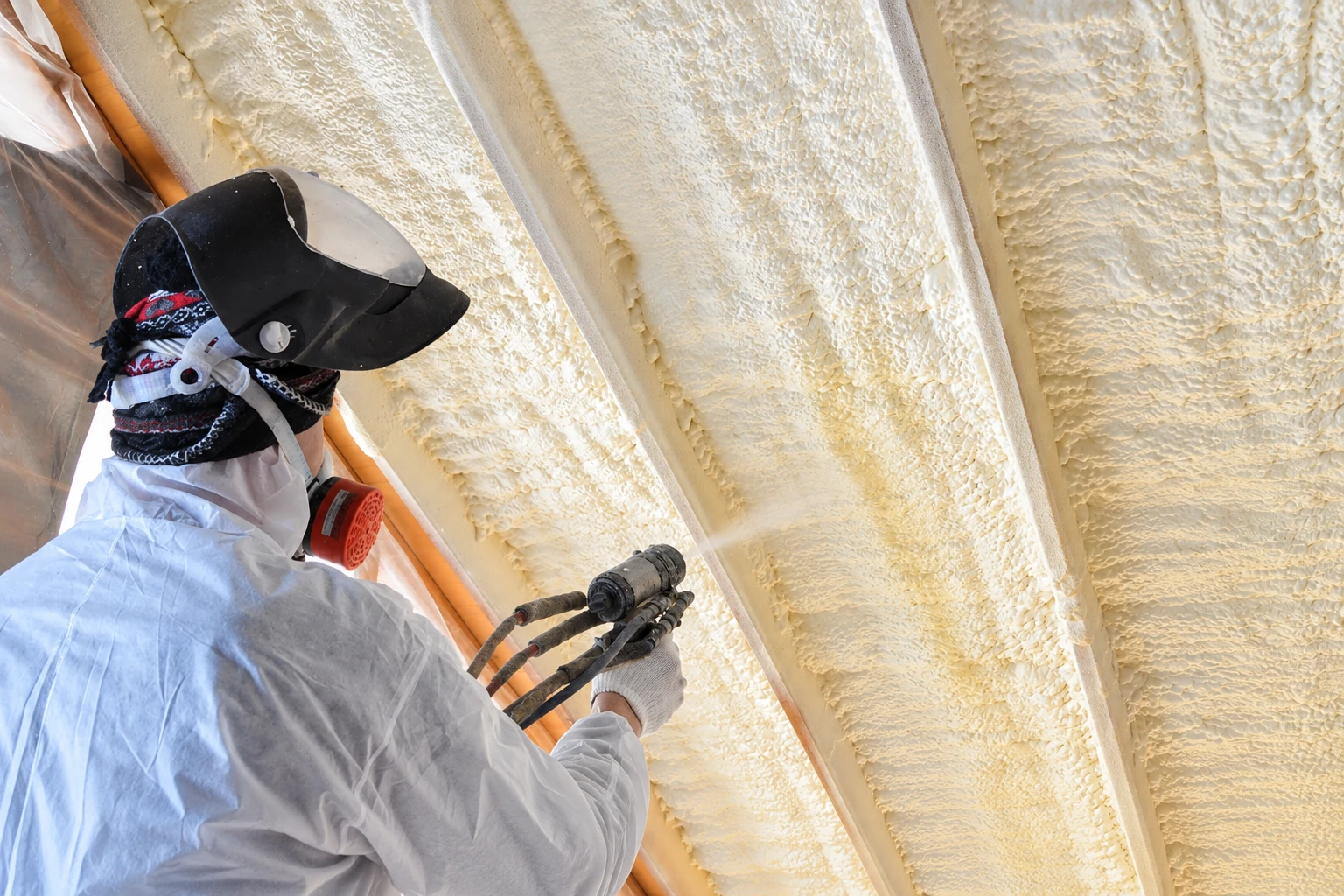 Worker in protective gear sprays foam insulation onto the underside of a roof between rafters.