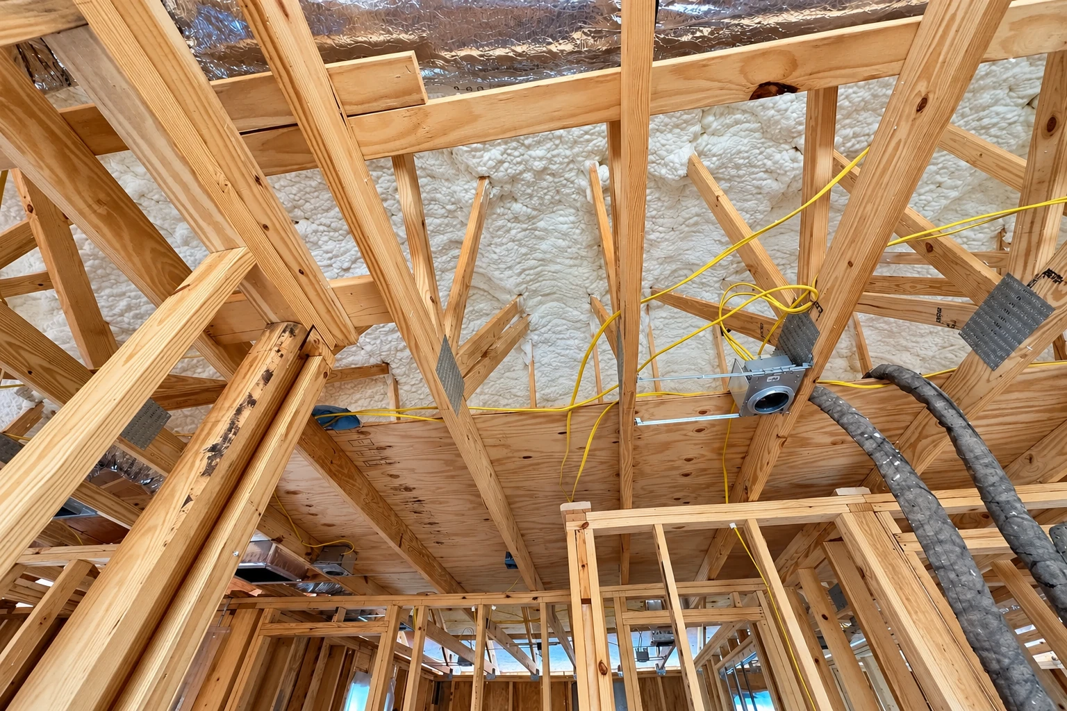Exposed wooden roof trusses with white spray foam coating the roof underside and flexible ducts and wiring between beams.