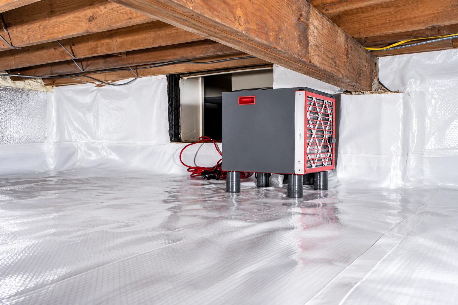 A crawl space has white vapor barrier sheeting across the floor and walls, surrounding a gray dehumidifier beneath wooden joists.