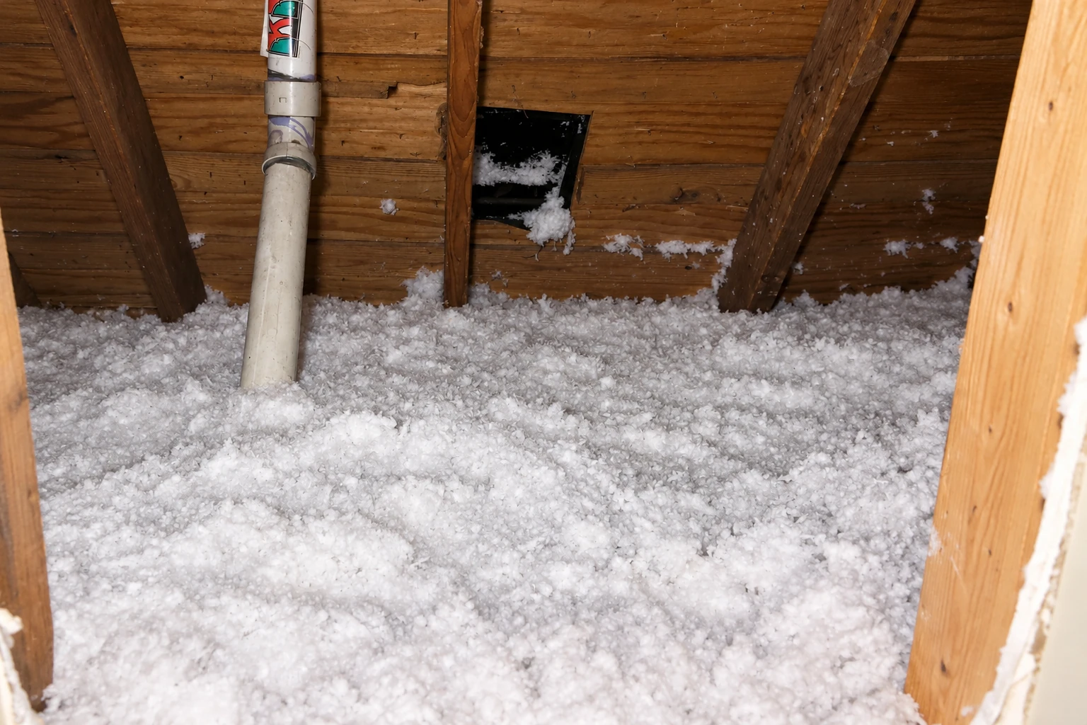 Loose-fill insulation blankets an attic floor around a white pipe, with exposed wooden roof framing above.
