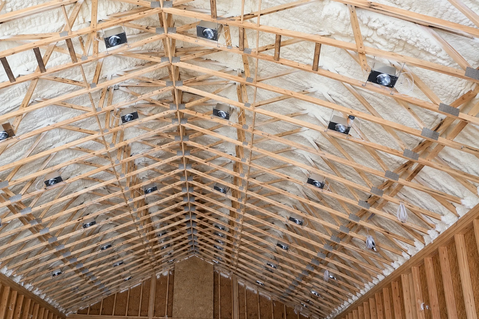 Spray foam insulation coats the roof underside between exposed wooden trusses, with several black fixtures mounted among the beams.
