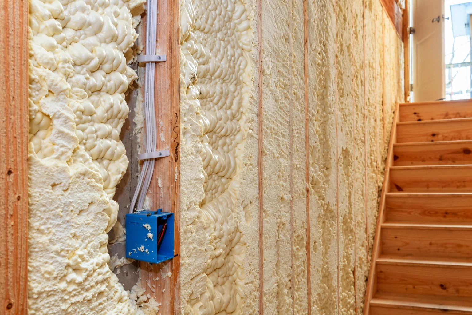 Wall studs packed with cream-colored spray foam insulation beside a wooden staircase.