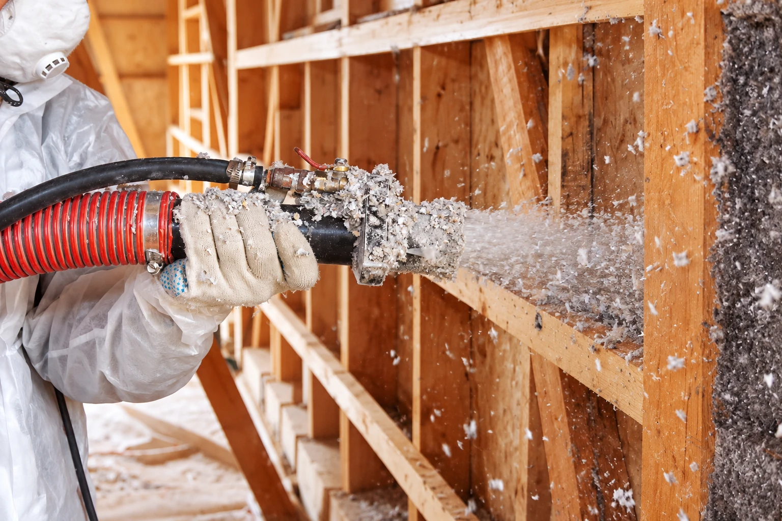 A worker in protective clothing directs a hose as loose-fill insulation is blown into an open wall cavity.